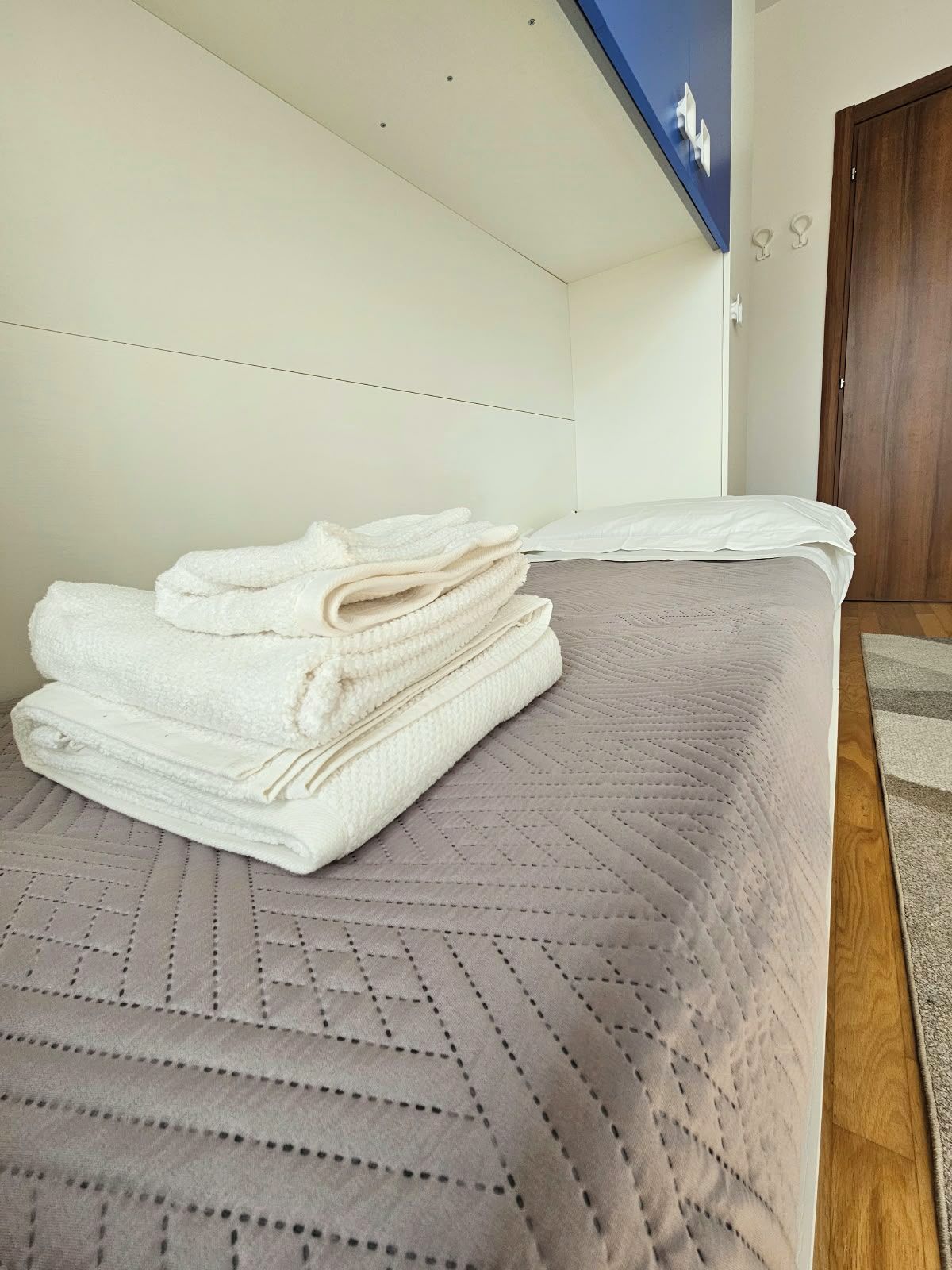 Ein ordentlich bezogenes Bett mit einem gefalteten weißen Handtuch darauf, einer hellen Bettdecke und einer Holztür im Hintergrund.