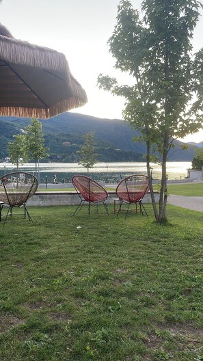 A grassy area with three wicker chairs facing a body of water, a thatched roof structure, and a tree.