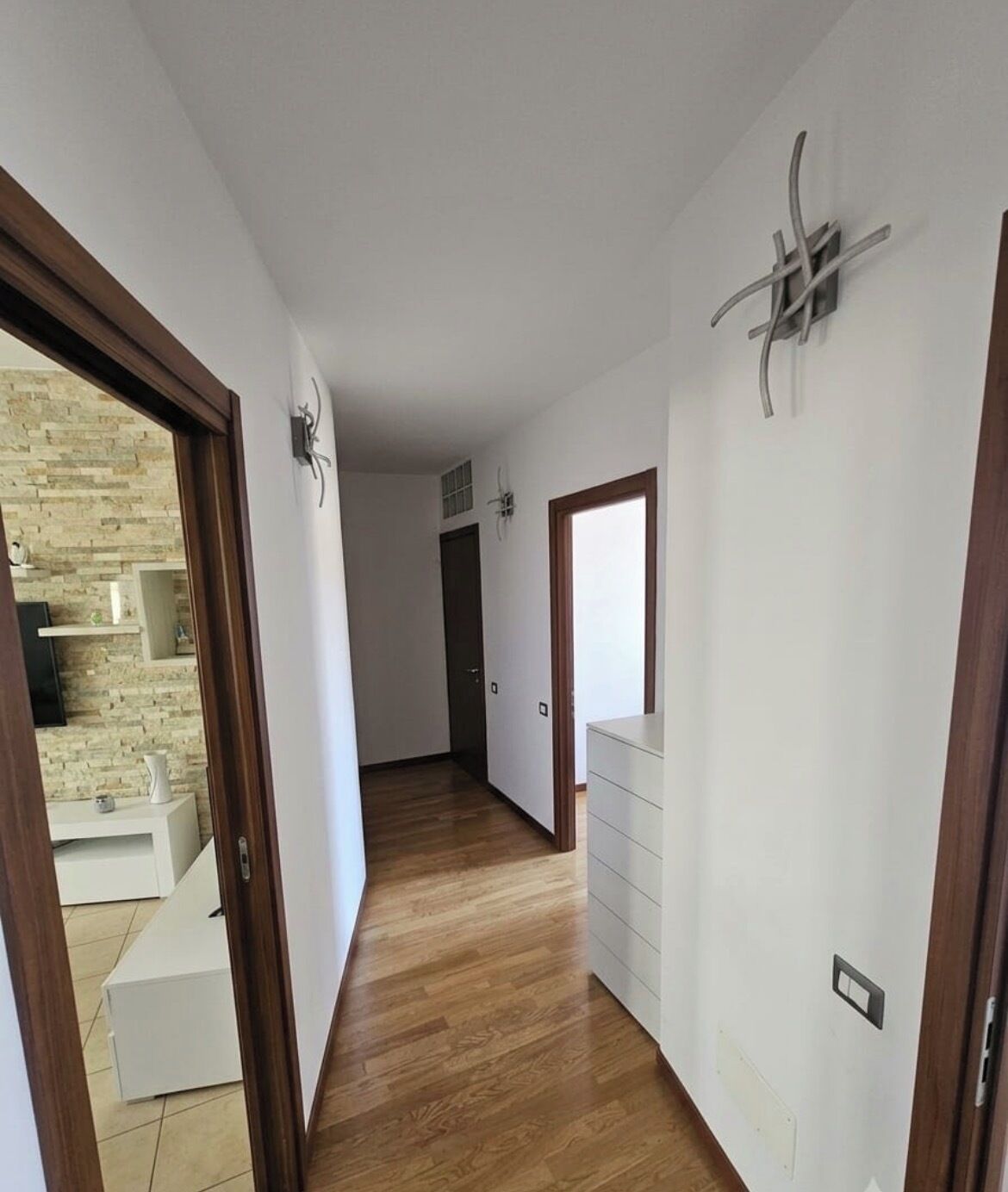 A hallway with wooden flooring, white walls, and a door leading to a room with a white dresser.