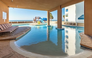 A pool area with lounge chairs and a slide, surrounded by a building with a view of the ocean.