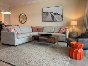 A living room with a sectional sofa, a coffee table, a patterned rug, a painting of a beach, and a lamp. - Phoenix Orange Beach 1202 (Orange Beach)