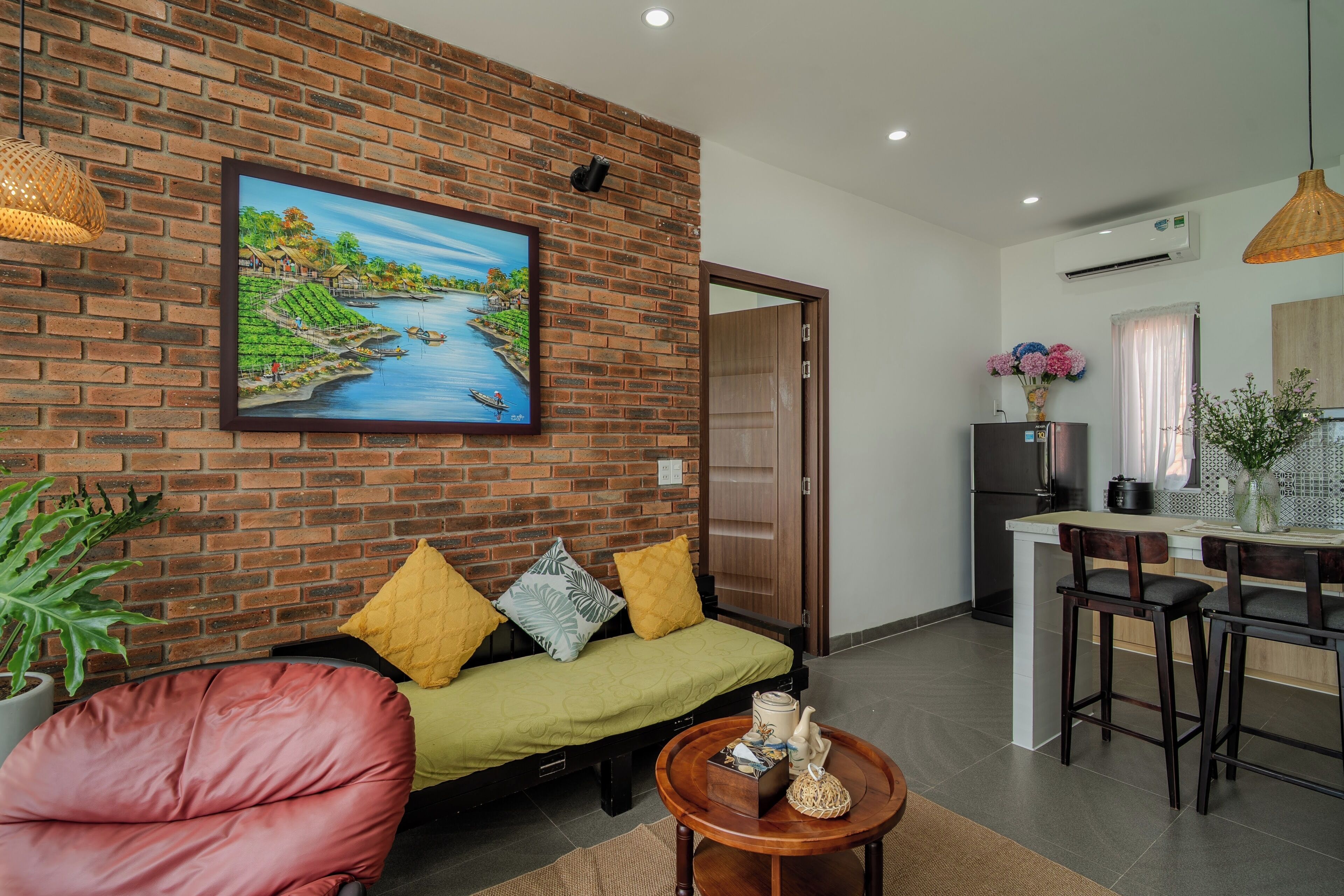 A living room with a brick wall, a sofa with cushions, a coffee table with decorative items, and a painting of a river scene.