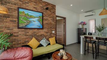 A living room with a brick wall, a sofa with cushions, a coffee table with decorative items, and a painting of a river scene.