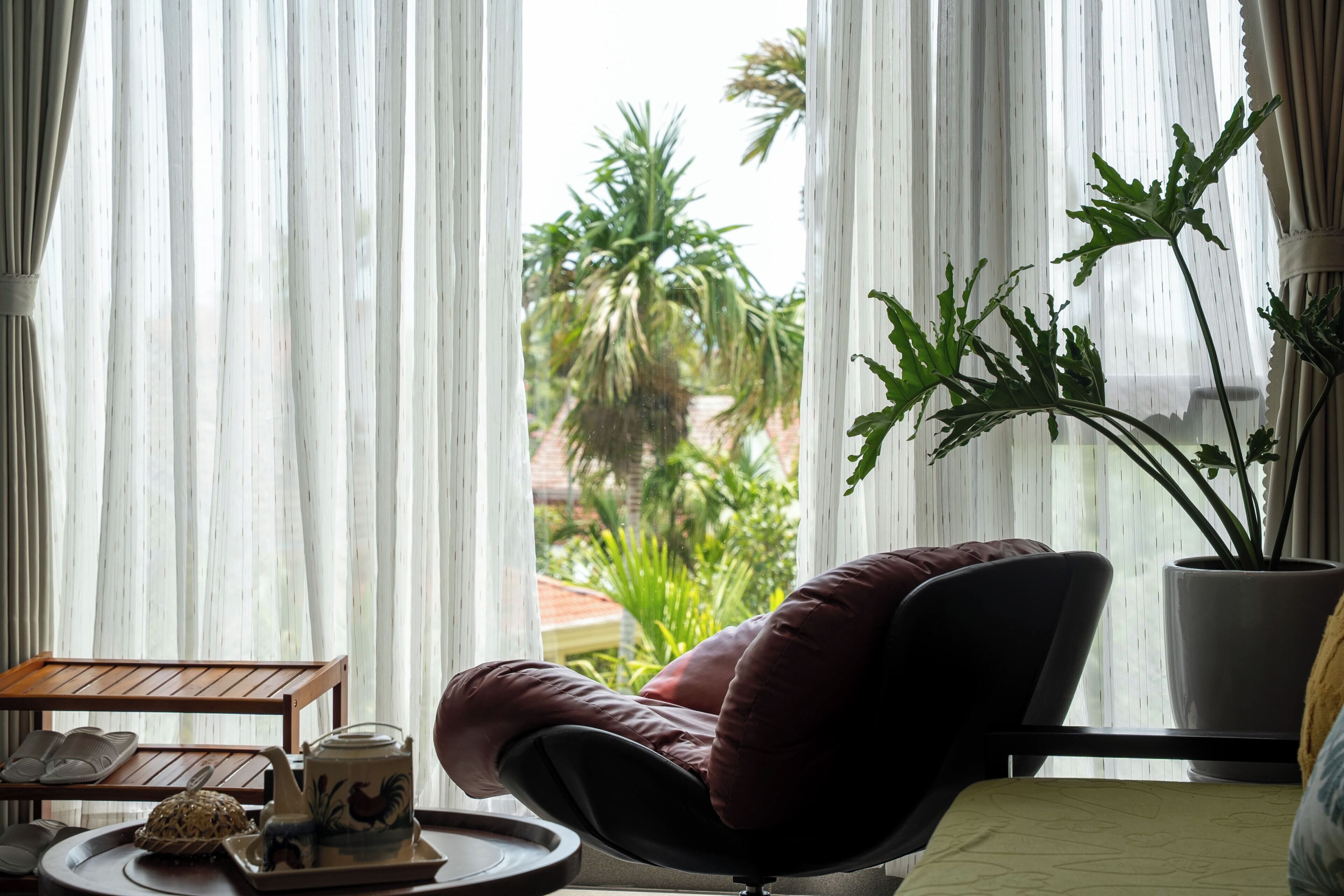 Una habitación con vistas a palmeras, una silla, una mesa con tetera y una planta en maceta.