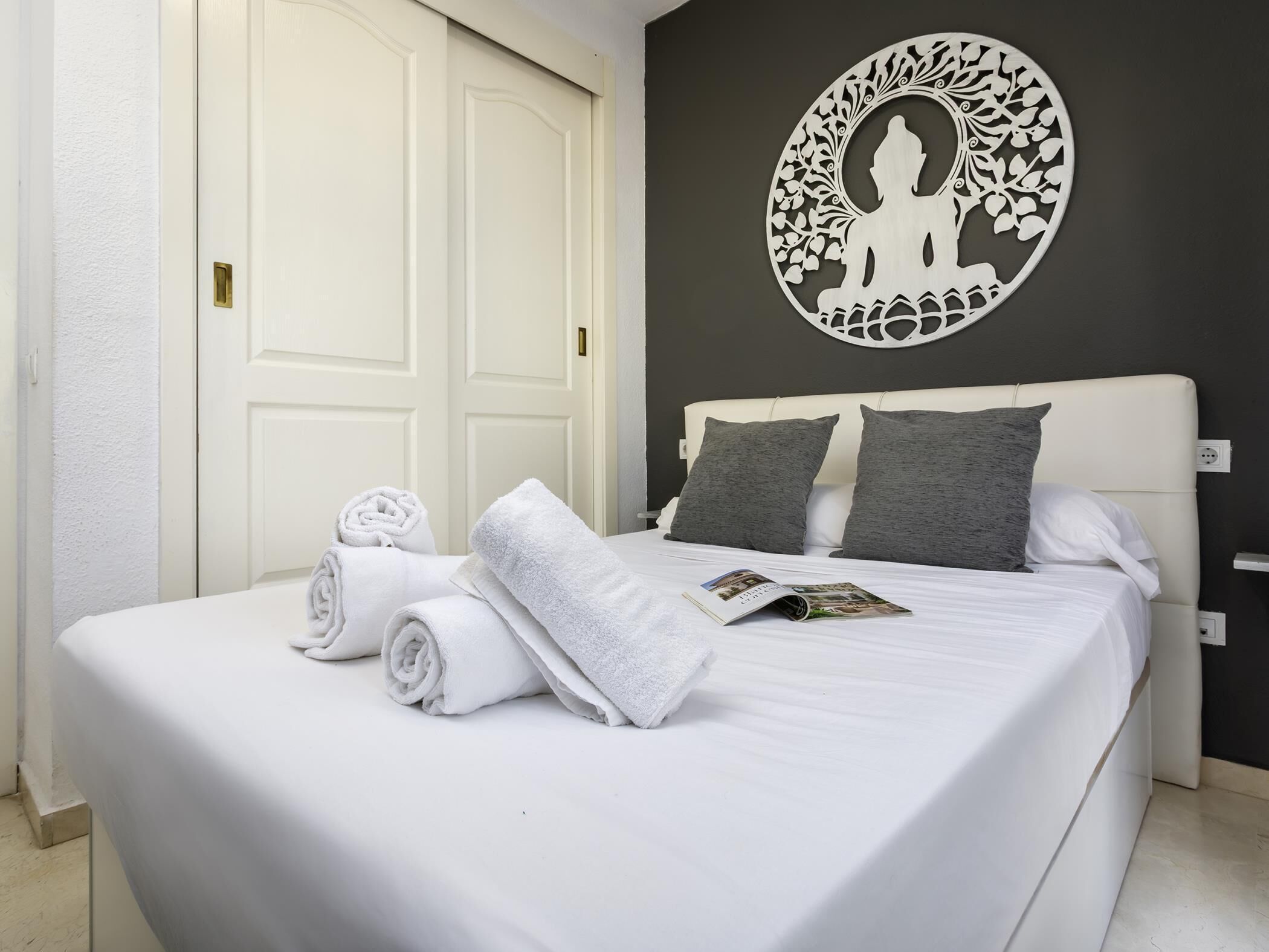 Hotellihuone, jossa on sänky, kaksi tyynyä, rullalle taitetut pyyhkeet, lehti ja koristeellinen seinäkoriste.