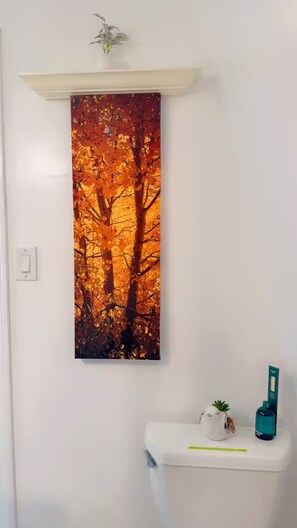 A bathroom with a wall-mounted painting of autumn leaves, a white toilet, and a small potted plant on the sink.
