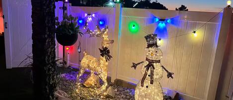 Un jardín con esculturas decorativas de un reno y un muñeco de nieve, rodeado por una valla de madera y luces de cadena.