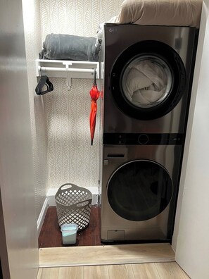 A compact laundry room with a front-loading washing machine and dryer, a drying rack, a basket, and a small trash bin. - The Manor Cottage (El Cajon)