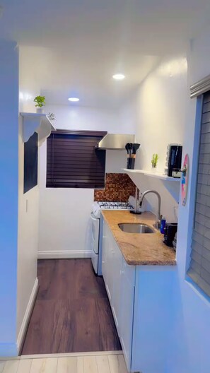 A compact kitchen with a sink, stove, and a window with blinds. - The Manor Cottage (El Cajon)