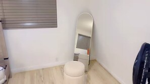 A small, clean room with a toilet, a mirror, a fan, and a window with blinds. - The Manor Cottage (El Cajon)