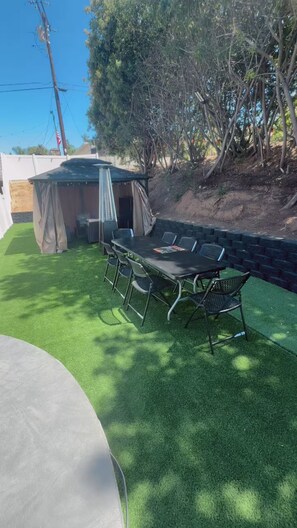 An outdoor patio with a table and chairs, surrounded by artificial grass and a covered seating area. - The Manor Cottage (El Cajon)