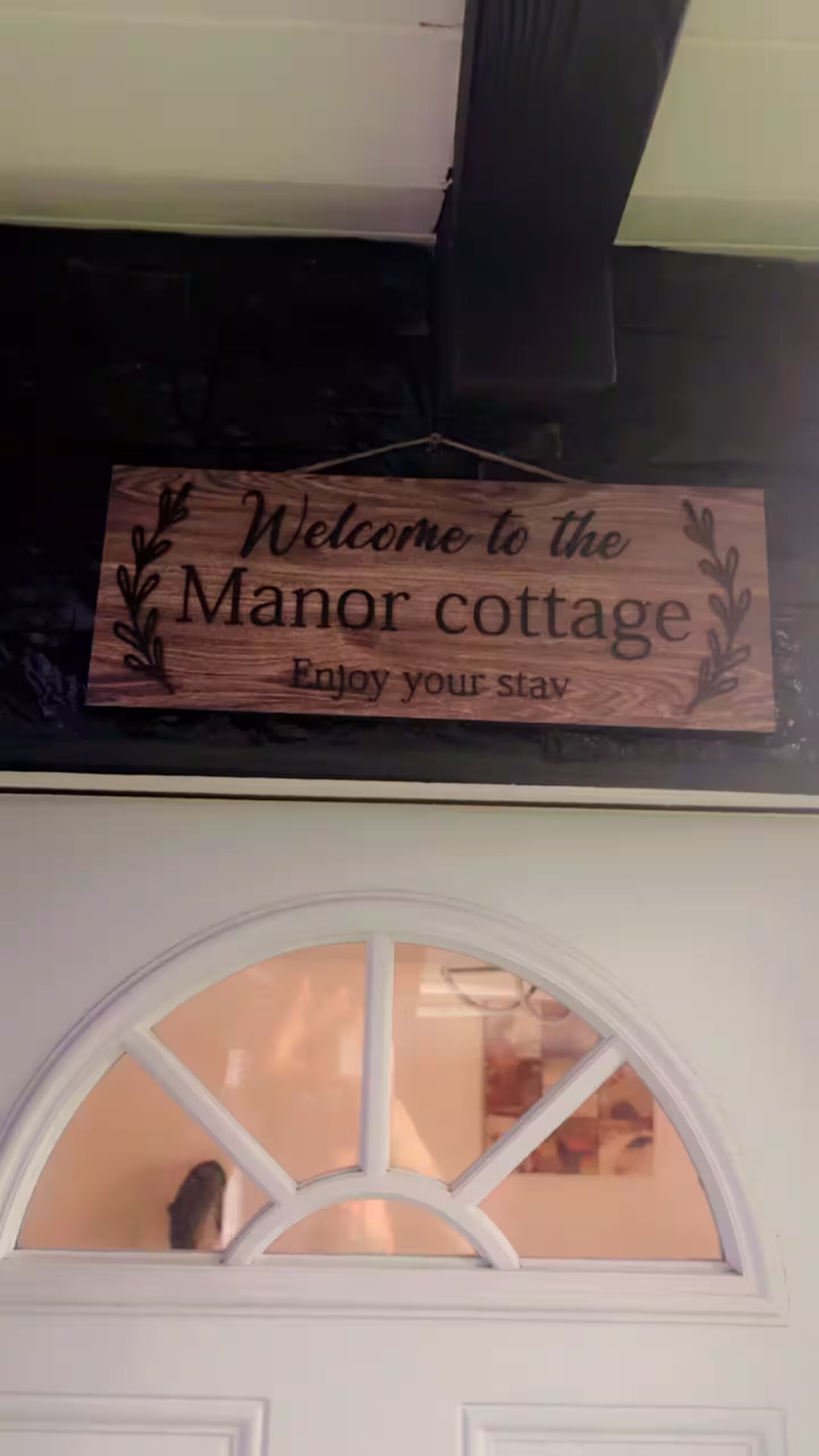 Un letrero de madera con el texto 'Welcome to the Manor cottage' y 'Enjoy your stay' sobre una puerta blanca con un ventanal semicircular.