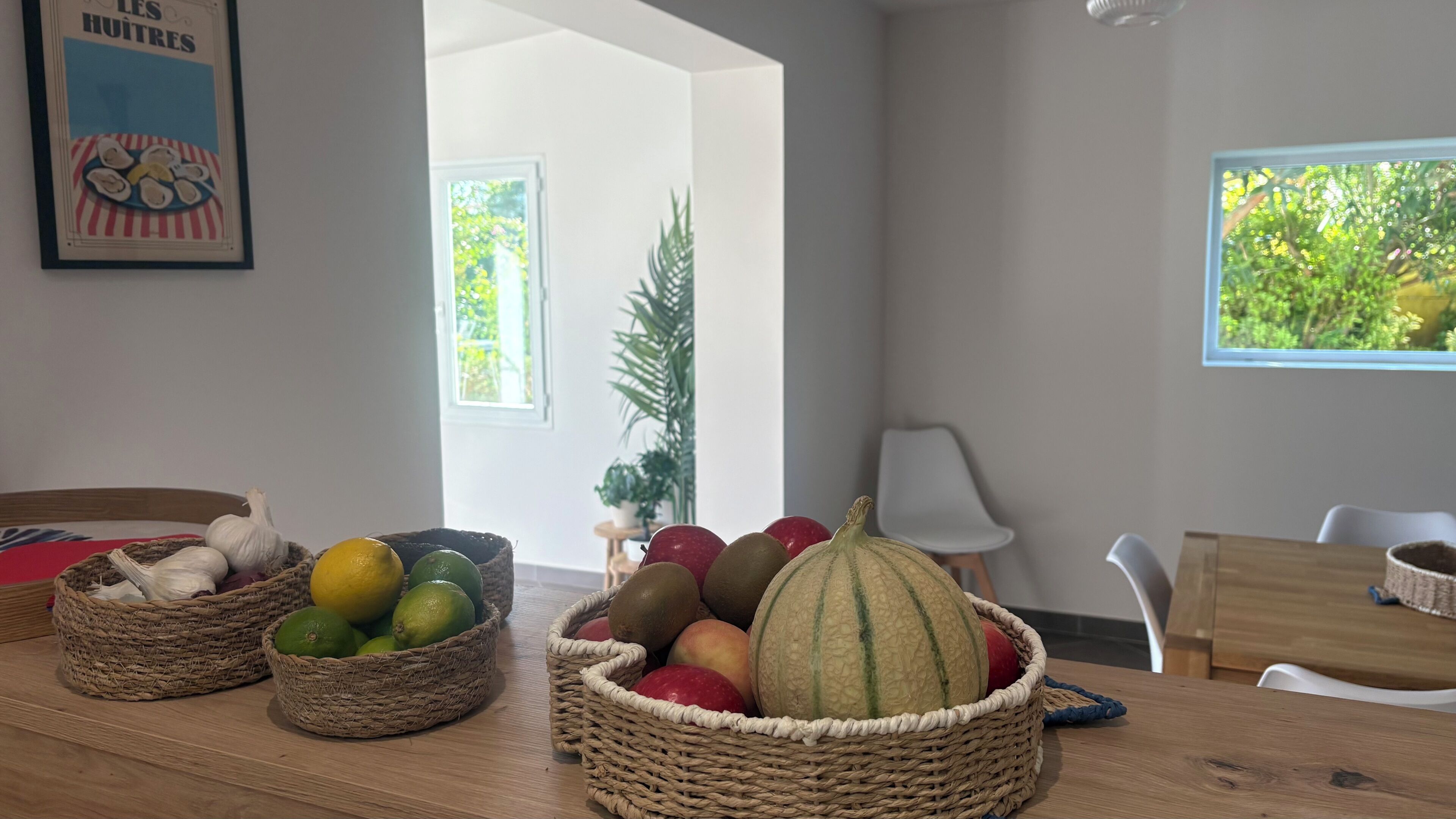 Un coin repas agrémenté de paniers en osier remplis de fruits et de légumes, d’une table en bois et d’une vue sur la verdure par une fenêtre.