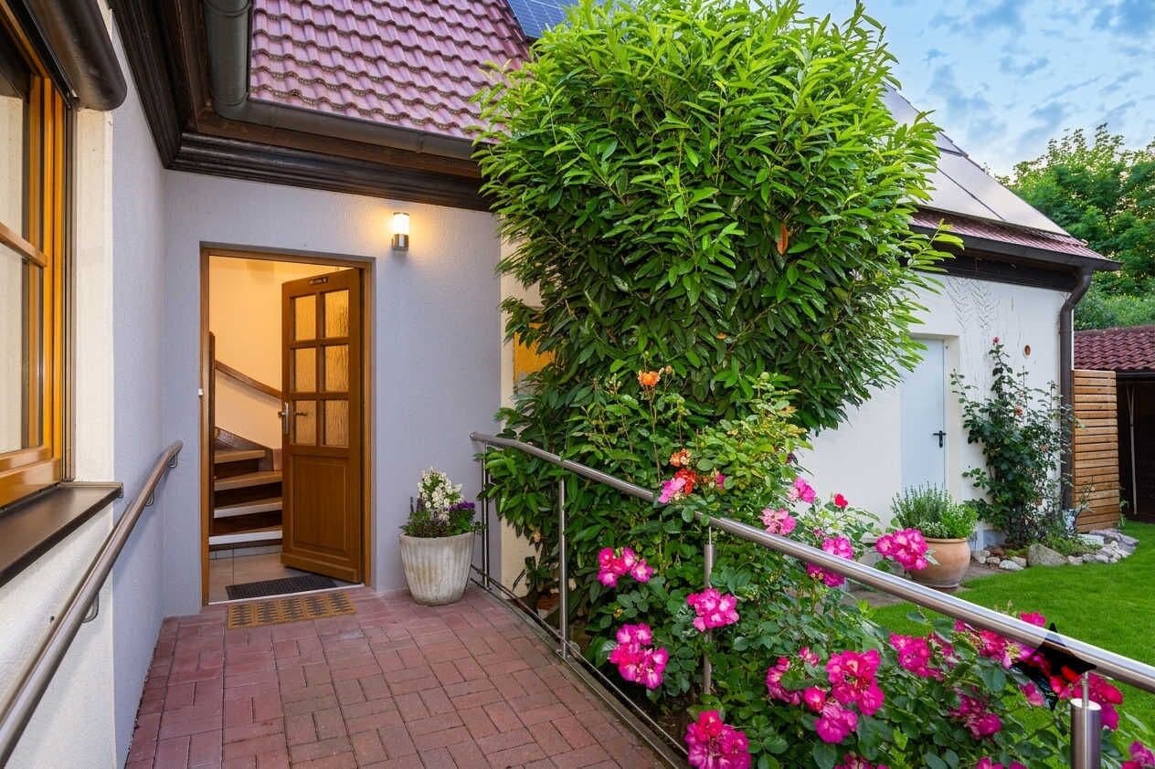 A house with a staircase leading to an interior, a garden with pink flowers, and a metal railing.