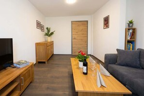 A modern living room with a flat-screen TV, wooden furniture, a dining table with a bottle and glasses, a sofa, and a bookshelf. - Wohlfühldomizil im Grünen (Leipheim)