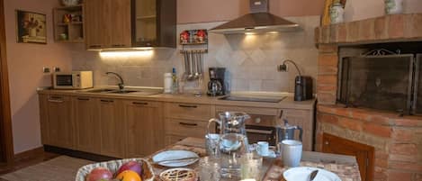 A kitchen with wooden cabinets, a stainless steel range hood, a stone fireplace, and a breakfast setup on the table.