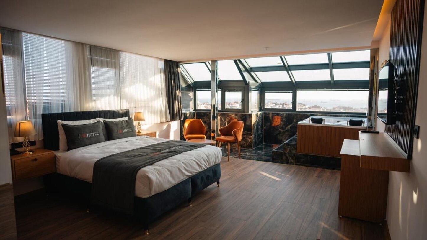 A hotel room with a large bed, a desk, two chairs, and a view through a skylight.