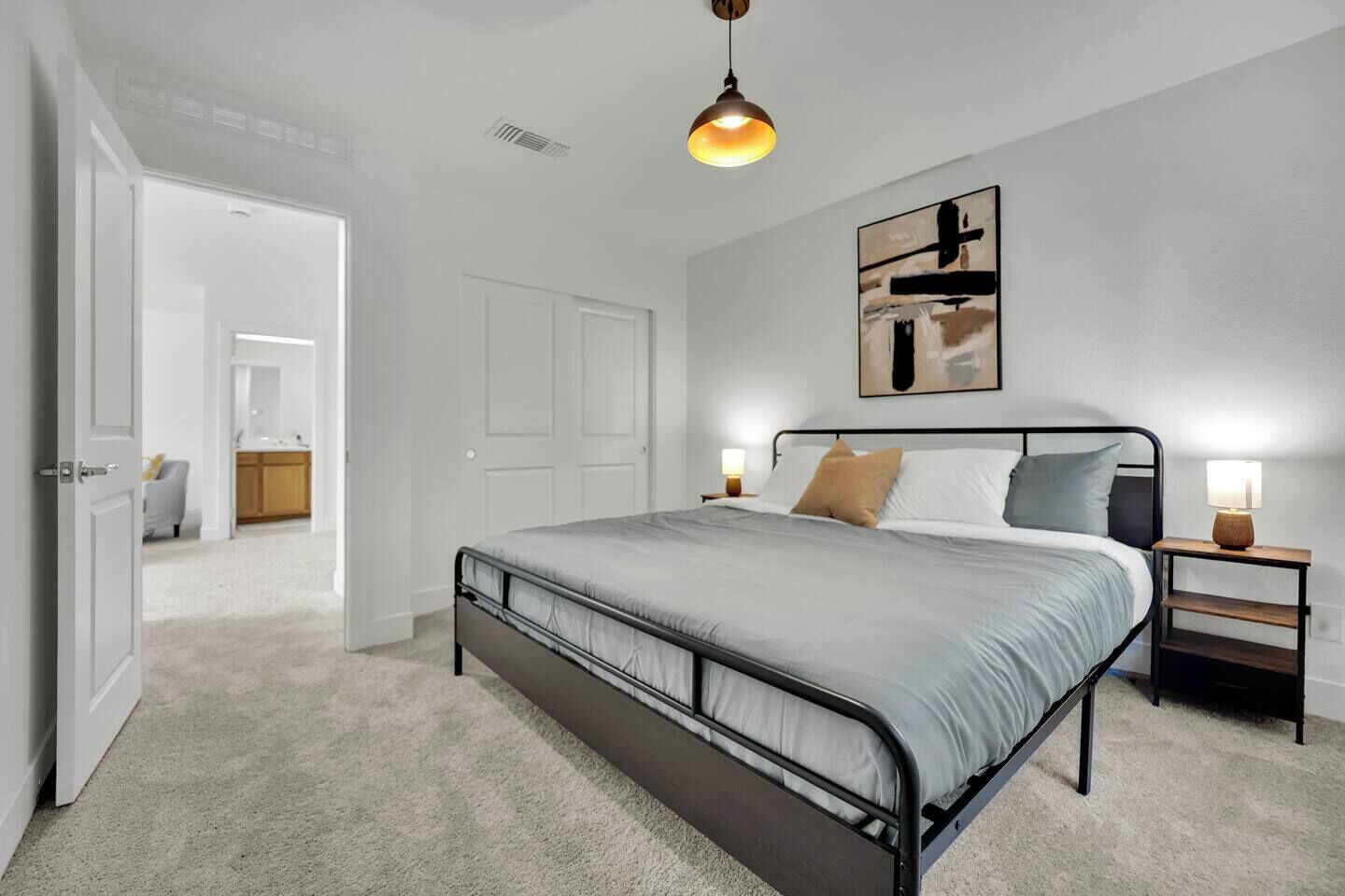 Um quarto com estrutura de cama de metal, colcha cinza, duas mesas de cabeceira com abajures e um quadro na parede.