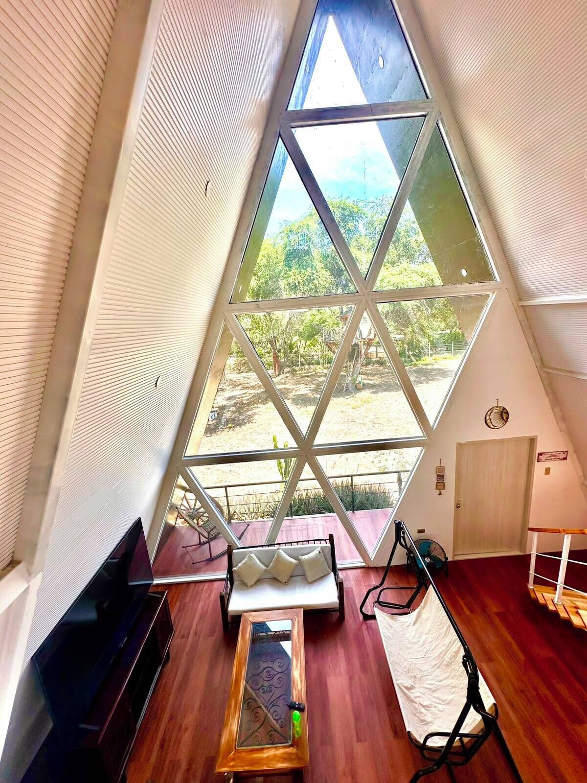 Una habitació amb sostre inclinat, un gran finestral triangular, una banca de fusta, una taula petita i una televisió.