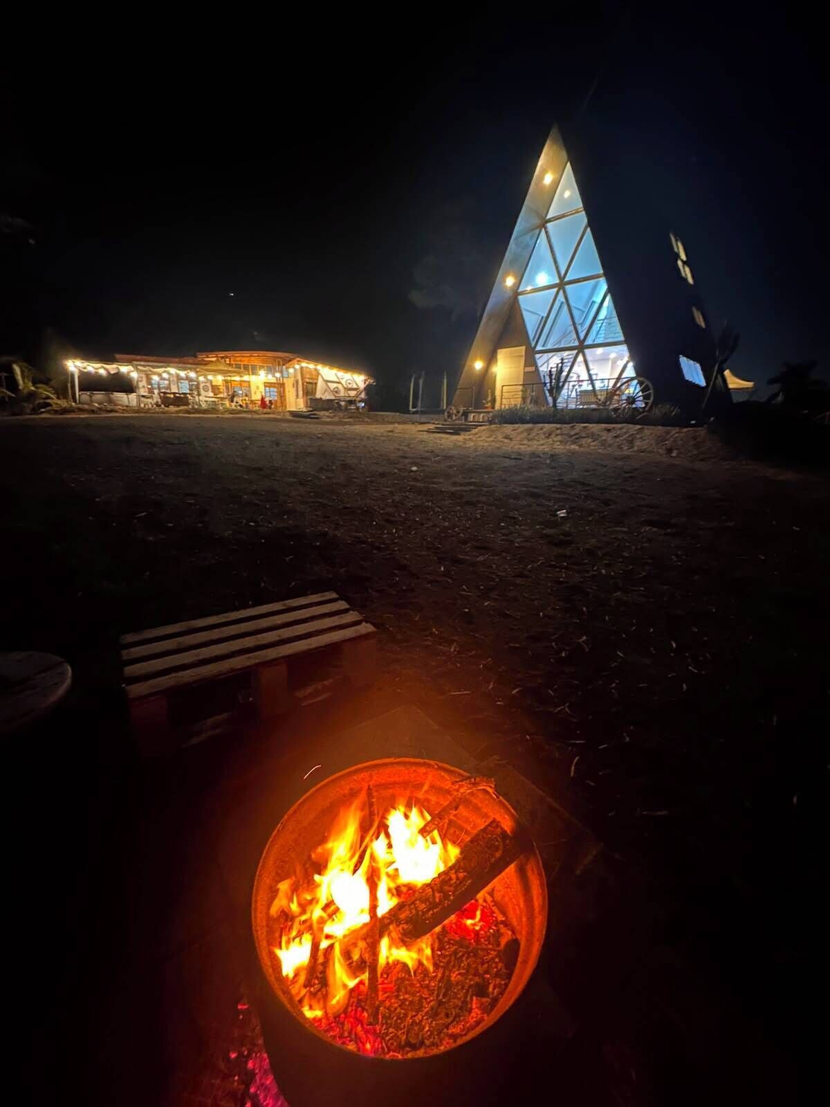 A campfire with logs and embers, a bench, and a building with a triangular glass facade at night.