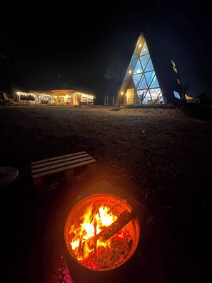 A campfire with logs and embers, a bench, and a building with a triangular glass facade at night.