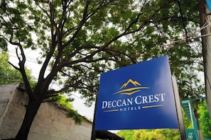 Un cartel de Deccan Crest Hotels con un logotipo de montaña, sobre un fondo que incluye un árbol y un edificio.