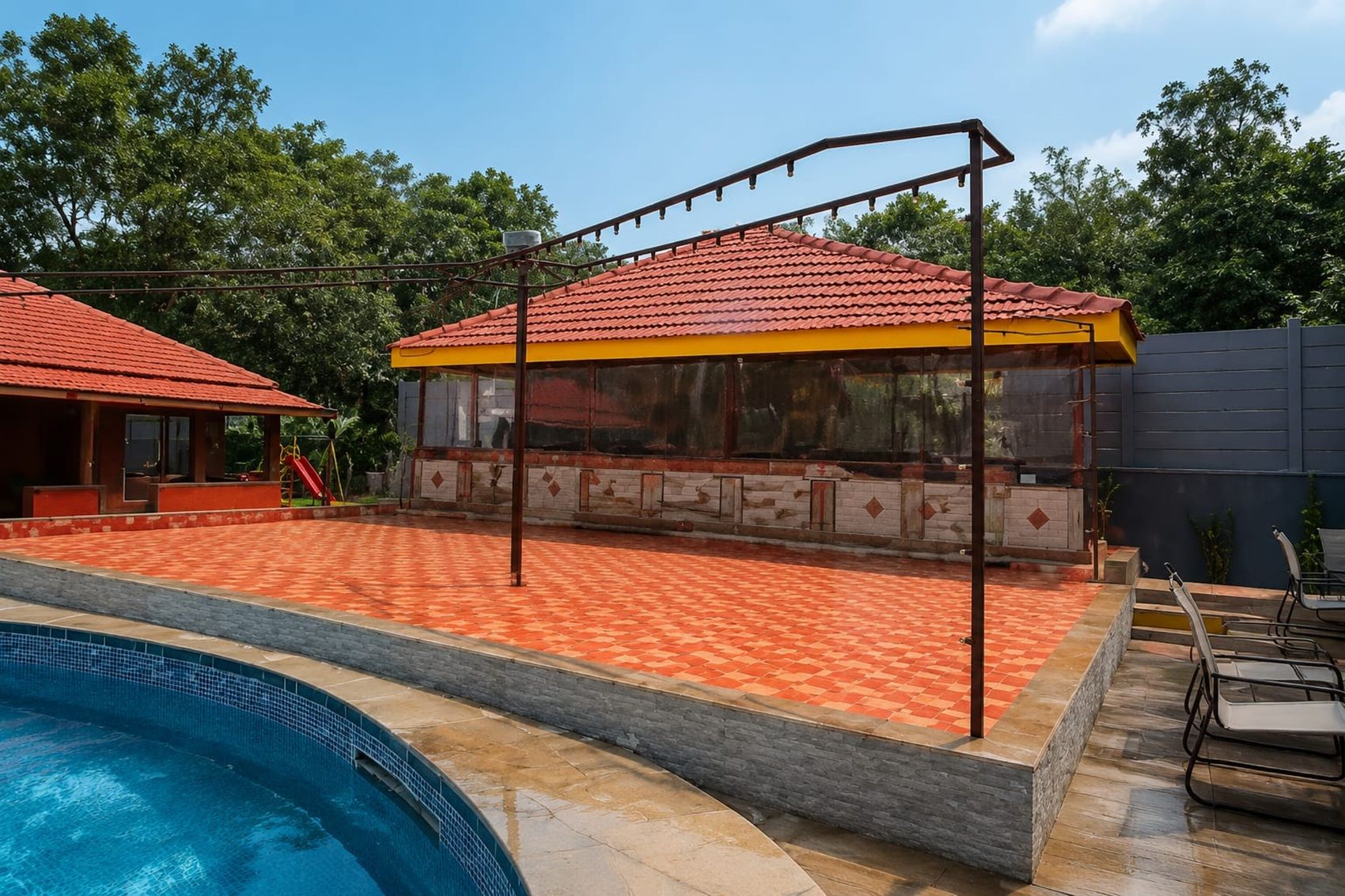 A swimming pool with a covered patio area, a children's play structure, and a building with a red-tiled roof.