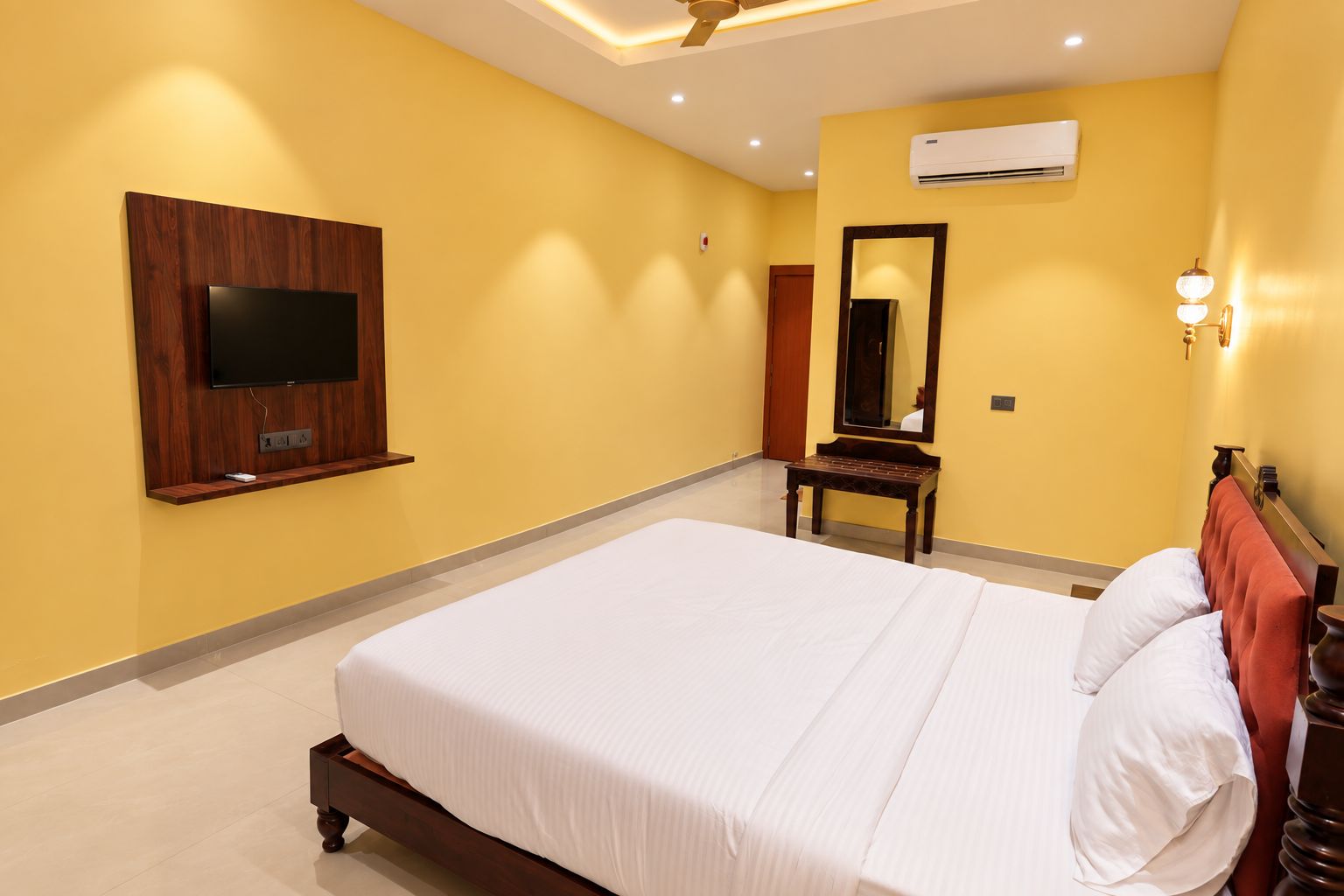 Ein Hotelzimmer mit einem großen Bett, einem flachbildfernseher an der Wand, einem kleinen Tisch, einem Spiegel und einem Deckenventilator.