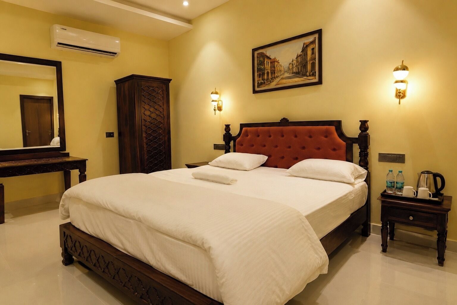 A hotel room with a large bed, a wooden headboard, a wall-mounted painting, a nightstand with a lamp, and an air conditioning unit.