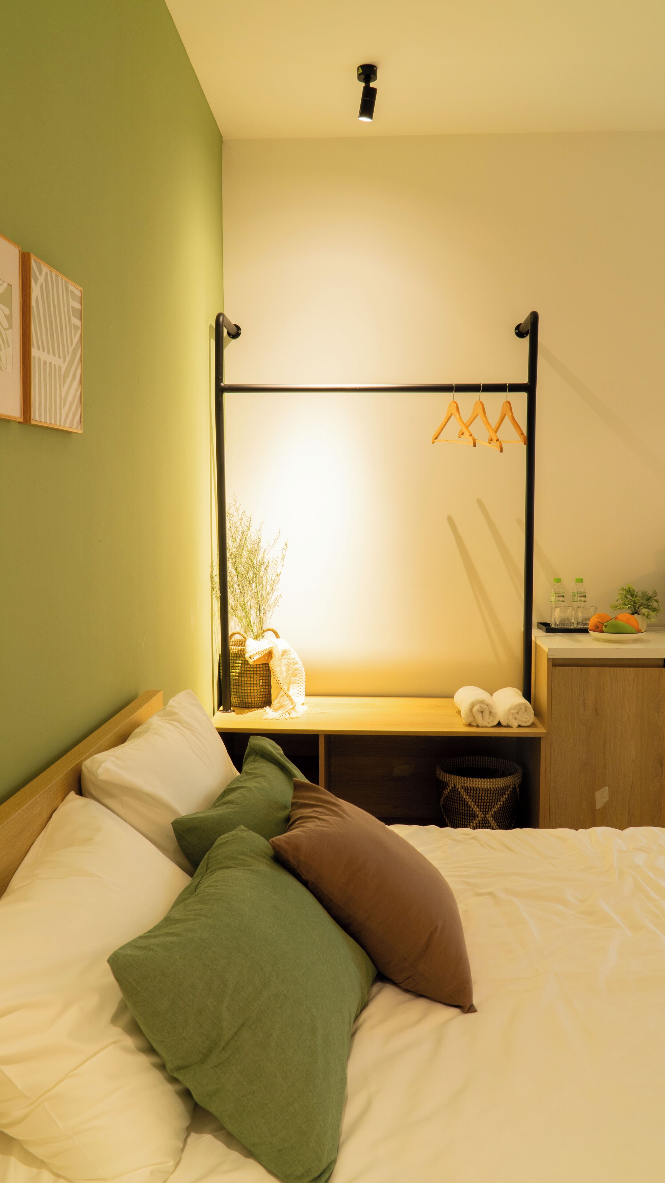 A room with a bed, a desk, a hanging light fixture, and a wall-mounted shelf with bottles and a plant.