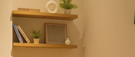 Een hoekkamer met een houten bankje, een geweven vloerkleed, een boekenplank met boeken en decoratieve objecten, en een wandplank met een plant en een ingelijst schilderij.
