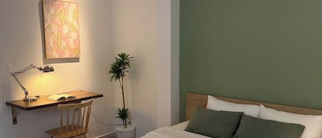 A bedroom with a bed, a desk with a chair, a lamp, and a plant.