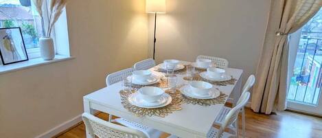 Un comedor con una mesa blanca dispuesta para cuatro, un reloj de pared y una ventana con vistas.