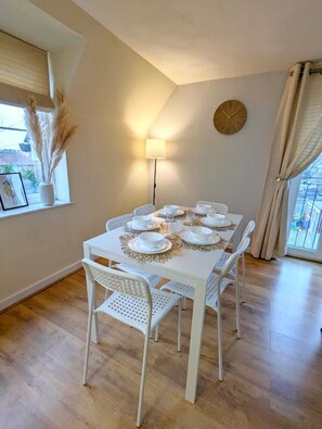 Een eetruimte met een witte tafel gedekt voor vier personen, een wandklok en een raam met uitzicht.