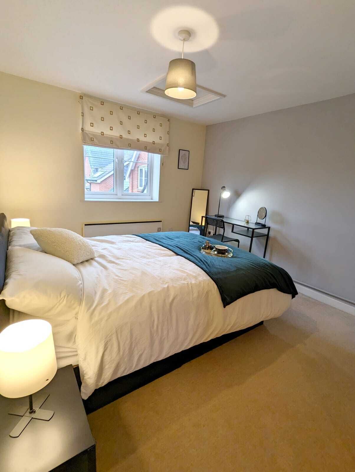 A bedroom with a bed, bedside table, lamp, desk with chair, and a window with a view of buildings.