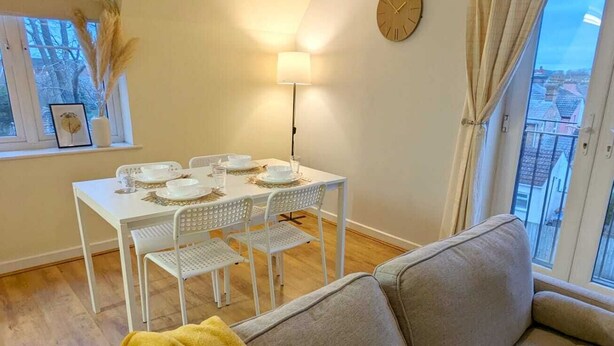 Un salón con sofá, una mesa de comedor con platos y vasos, una lámpara, un reloj y una ventana con cortinas.