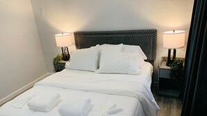 A neatly made bed with white linens and towels, a dark headboard, two bedside lamps, and a nightstand with a plant.