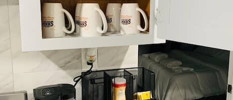 A kitchen with white mugs on shelves, a coffee maker, a black mesh organizer with items, and a bed in the background.