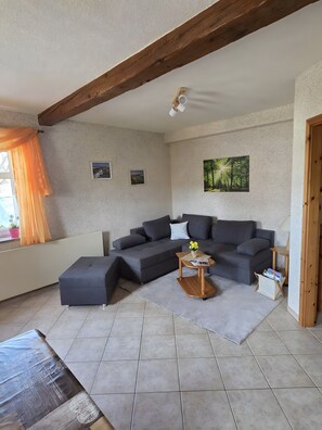 Ein Wohnzimmer mit einem Ecksofa, einem kleinen Holztisch und einem Fenster mit Vorhängen.