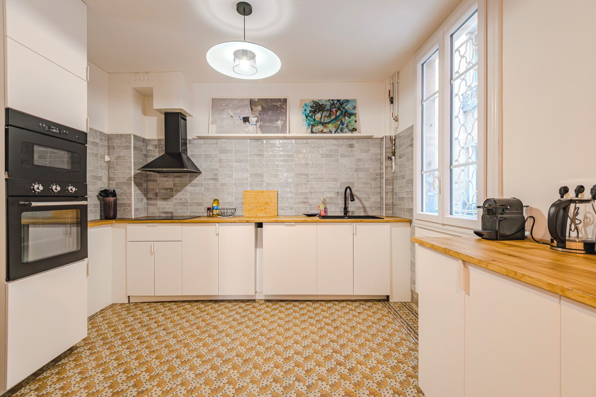 Uma cozinha com duas fornos, um exaustor preto, bancada de madeira, pia e janela com detalhes decorativos.