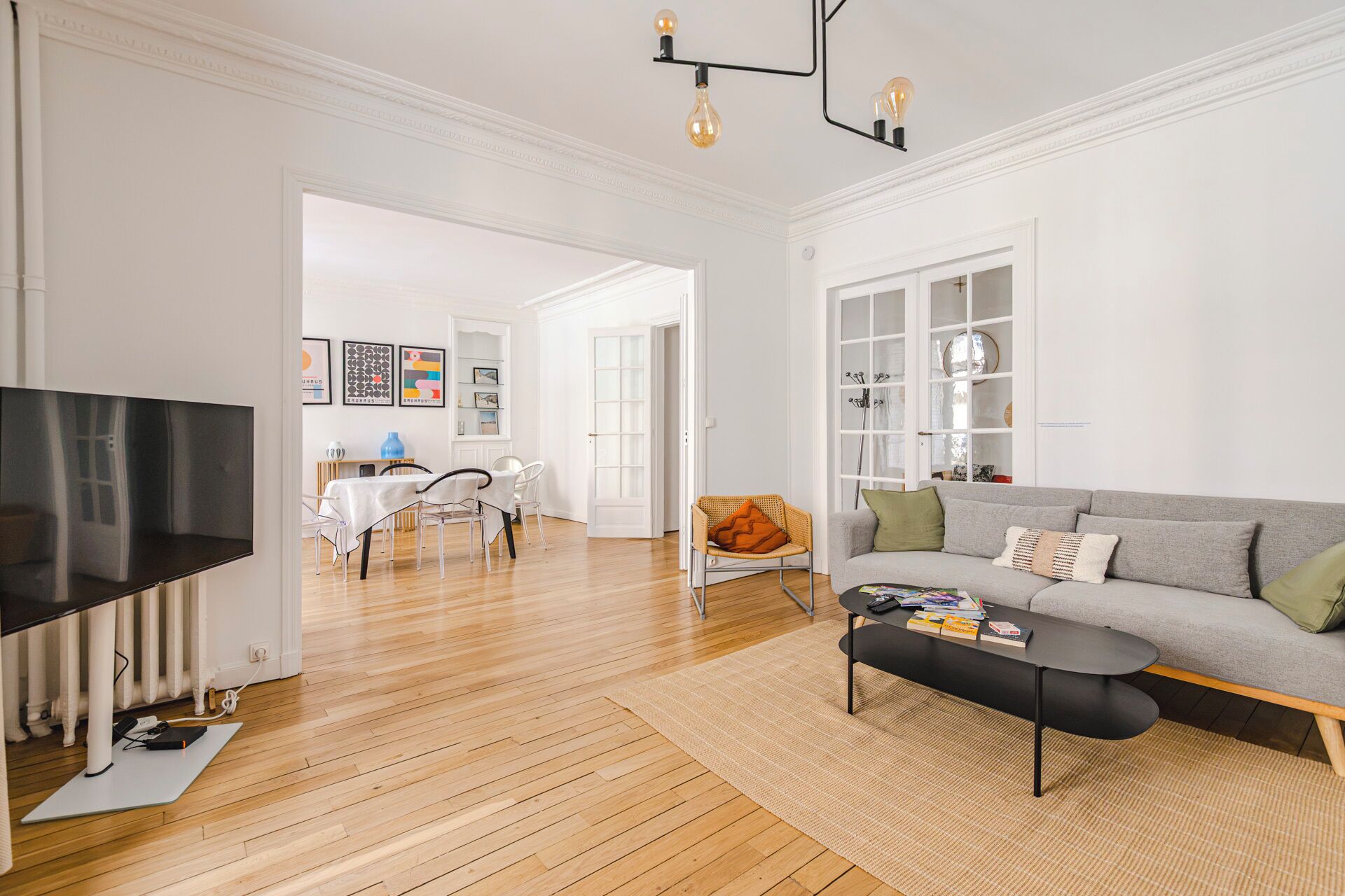 A modern living room with a sofa, coffee table, and a television. There is a dining area with chairs and a table, and a bookshelf filled with books and decorative items.