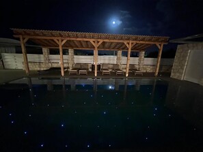 Ein Schwimmbecken mit Holzdeck und Liegestühlen, unter einem überdachten Bereich mit einer Steinwand im Hintergrund.