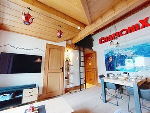 A room with a dining table, chairs, a TV, a wooden door, and a wall with a mountain painting. - Ginabelle 1 by Interhome (Chamonix)