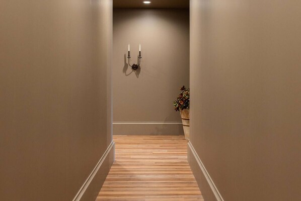 A narrow hallway with wooden flooring and beige walls, leading to a room with a potted plant.