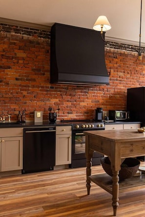 A kitchen with a brick wall, wooden flooring, a black stove, and a table with a lamp. - Historic King Suite | Downtown Snohomish Views (Snohomish)