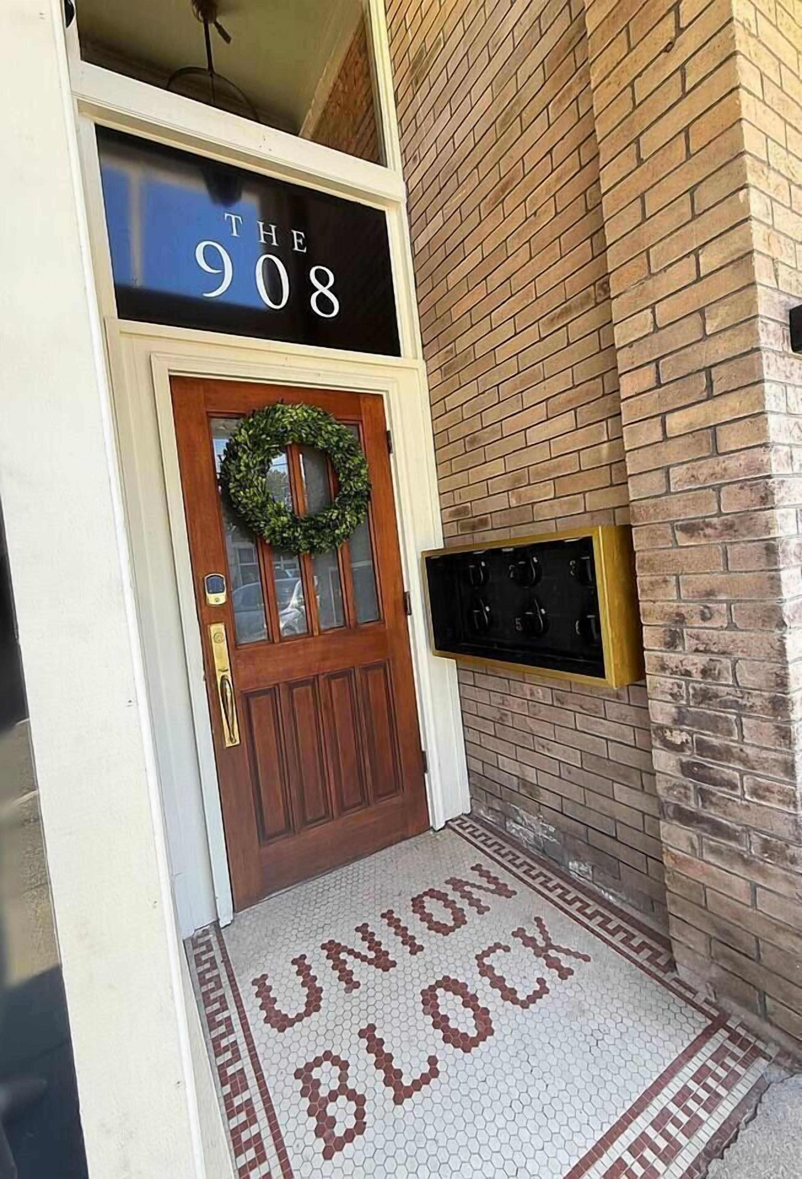 Une porte en bois ornée d’une guirlande, une enseigne au-dessus indiquant « THE 908 », et un paillasson « UNION BLOCK » à l’entrée.