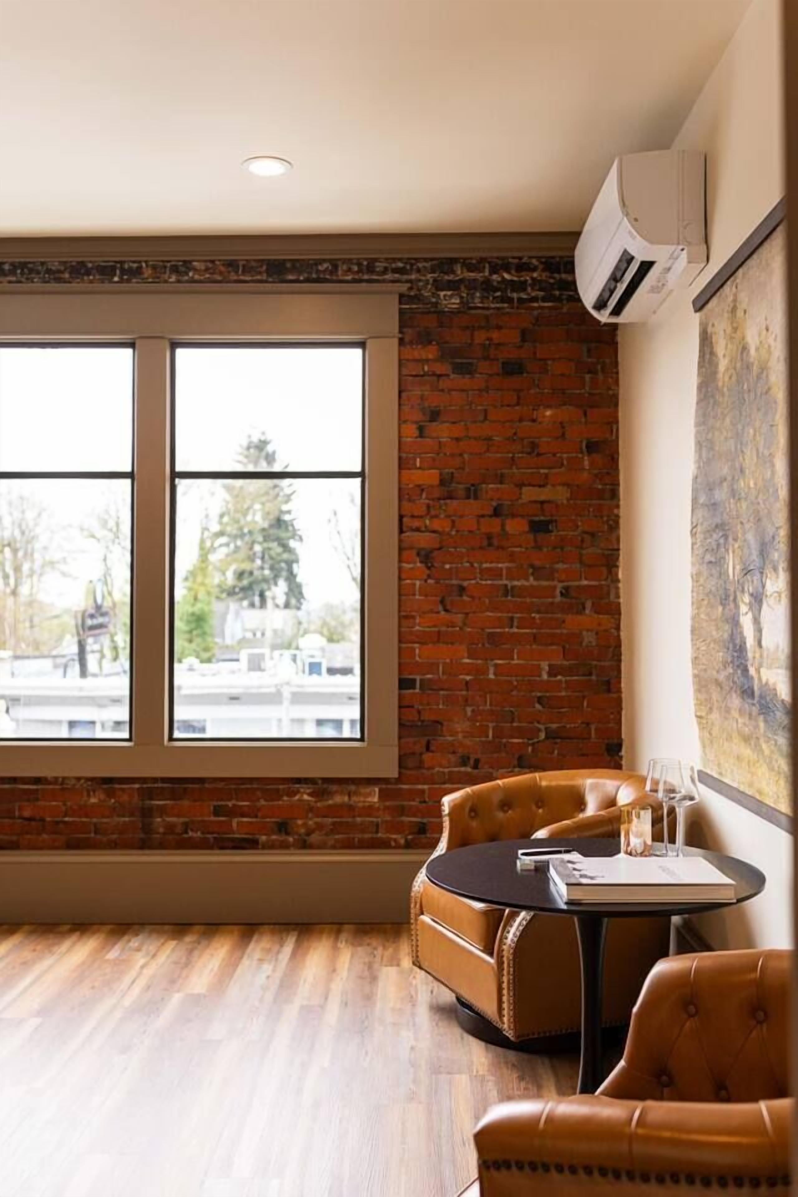 A room with a brick wall, a large window, a round table with chairs, and an air conditioning unit.