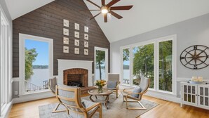 A spacious room with a high ceiling, a fireplace, a dining area with chairs, and large windows offering a view of the outdoors. - Lakefront + Private Dock | Hot Tub + Pool Table + Cabana | Point of View (Mooresville)