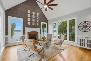 A spacious room with a high ceiling, a fireplace, a dining area with chairs, and large windows offering a view of the outdoors. - Lakefront + Private Dock | Hot Tub + Pool Table + Cabana | Point of View (Mooresville)