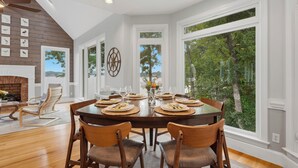 A dining room with a large table set for six, surrounded by wooden chairs, large windows with a view of trees, and a fireplace with a brick surround. - Lakefront + Private Dock | Hot Tub + Pool Table + Cabana | Point of View (Mooresville)
